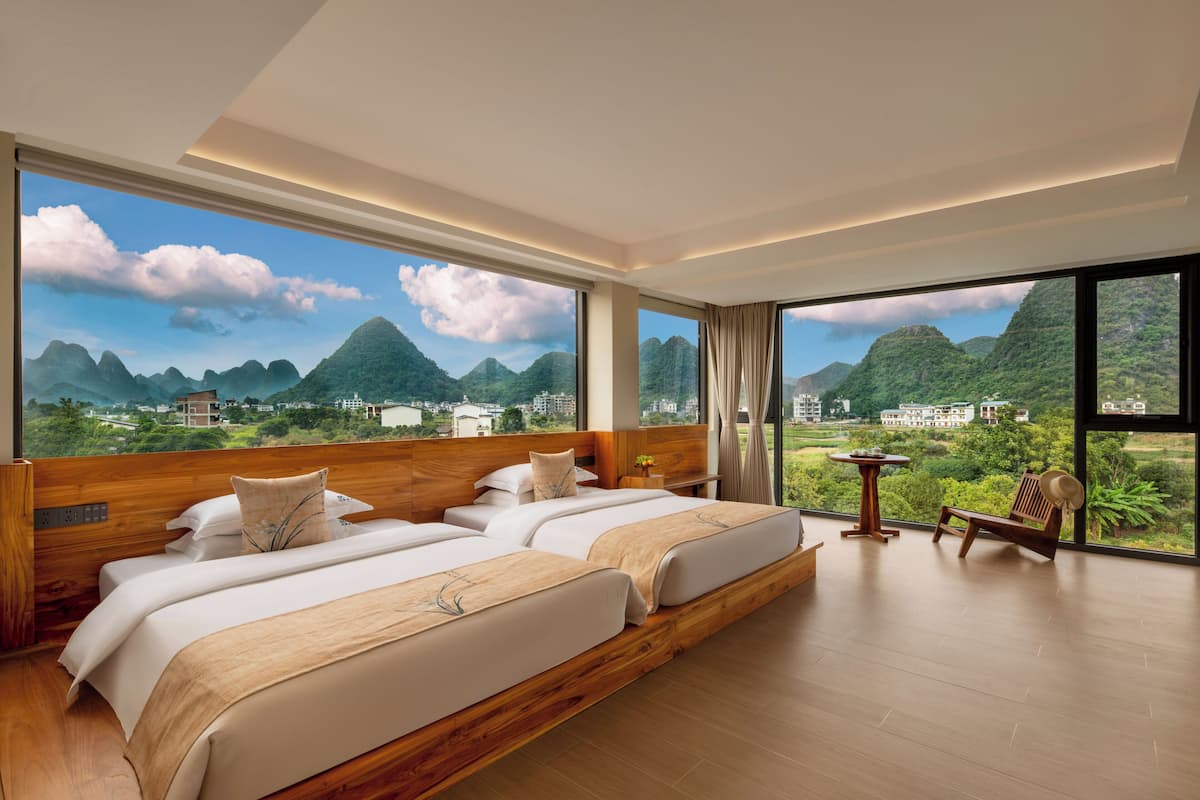 Habitación de hotel con un ventanal grande, dos camas, suelo de madera y vistas a montañas.