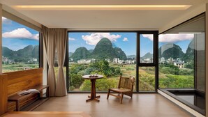 Una habitación con un ventanal grande, un banco de madera, una mesa pequeña con tazas y vistas a colinas verdes y edificios.