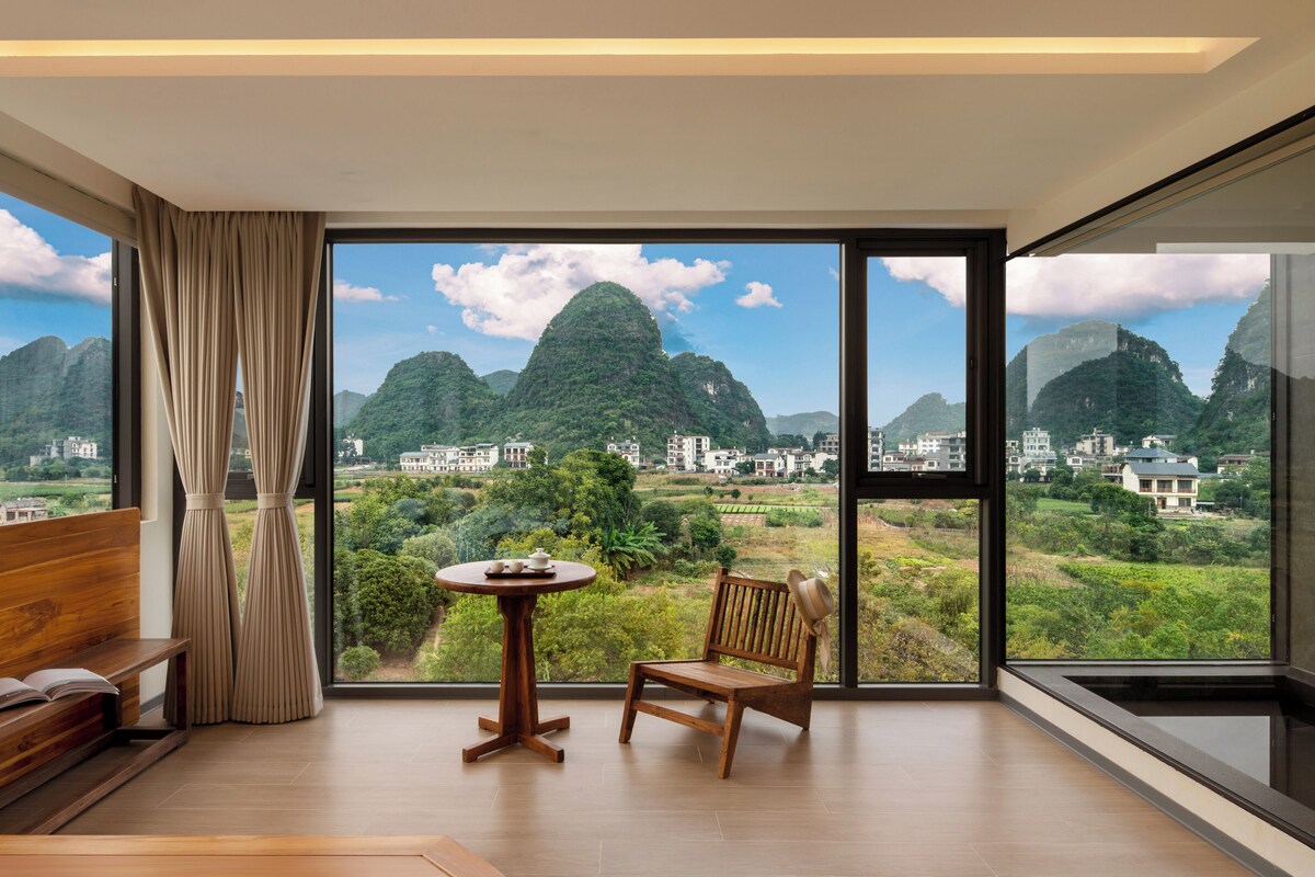 A room with a large window, a wooden bench, a small table with cups, and a view of green hills and buildings.