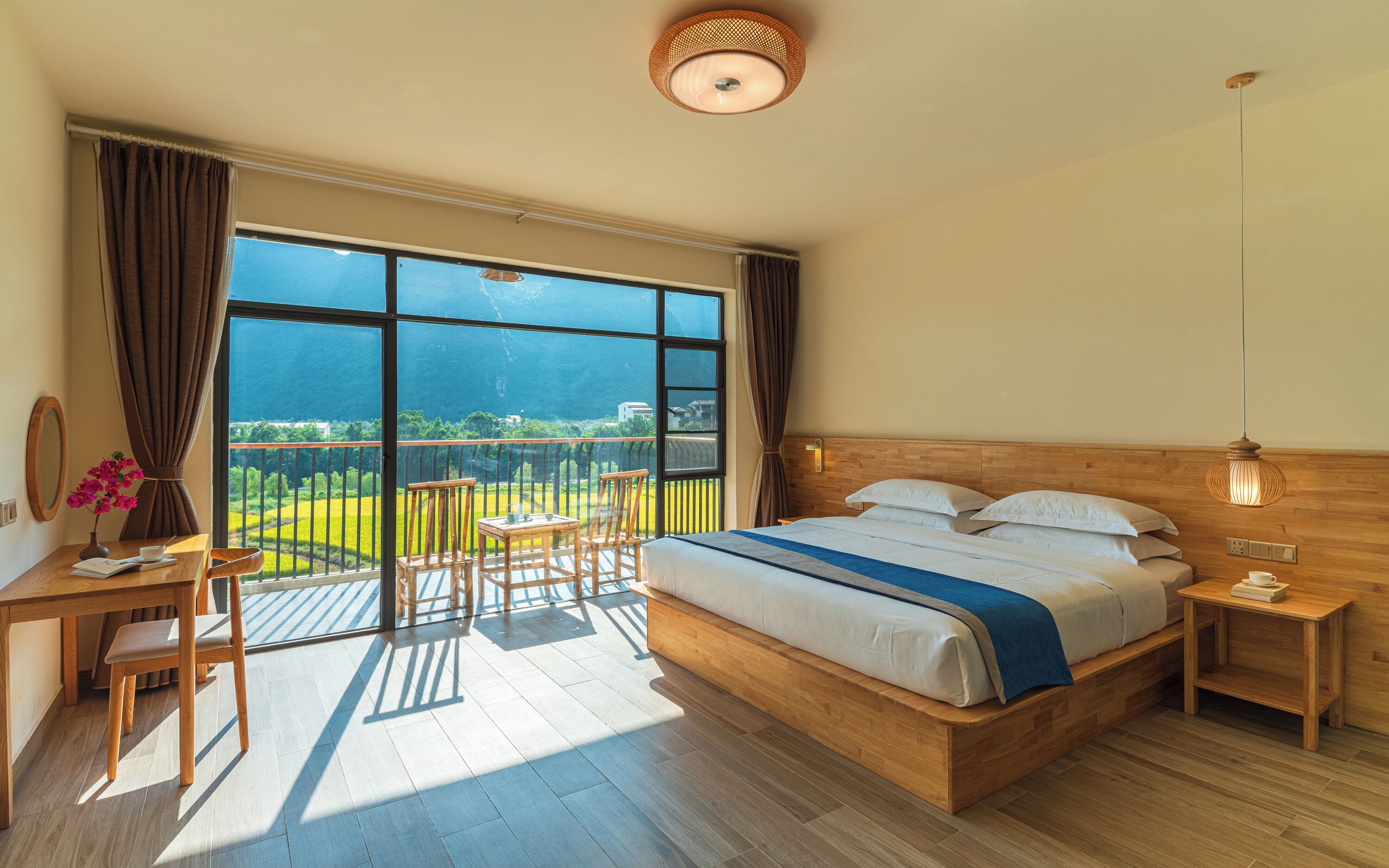 Een slaapkamer met een groot raam, een houten bed, een klein tafeltje, een stoel en een nachtkastje.
