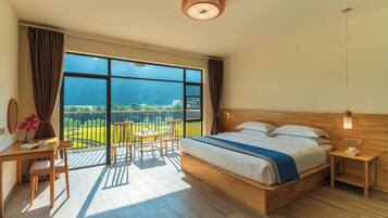 Een slaapkamer met een groot raam, een houten bed, een klein tafeltje, een stoel en een nachtkastje.