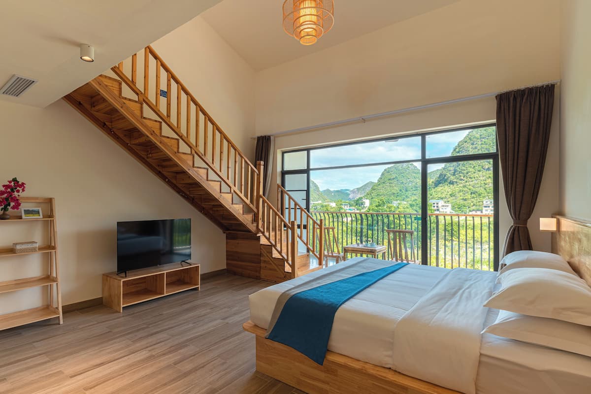 Un dormitorio con una escalera de madera, una cama con ropa de cama blanca, un televisor sobre un soporte de madera y un ventanal con vistas a la vegetación.