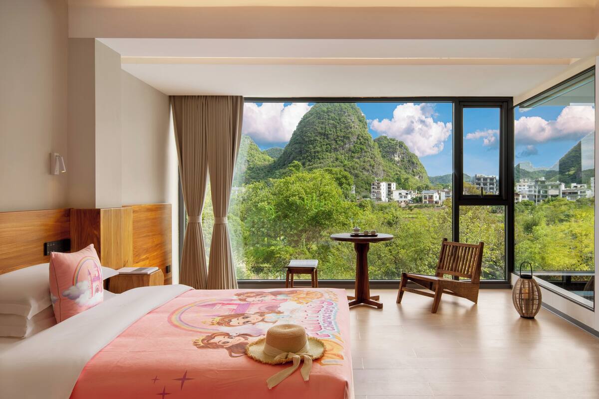 Habitación de hotel con cama, una silla, una mesita y vistas a un paisaje montañoso.