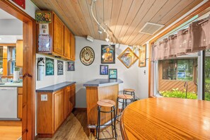 A kitchen with wooden cabinets, a bar area with stools, and various framed pictures on the wall. - Stealth Cottage – Vintage Stay Across from Paw Paw Lake (Coloma)