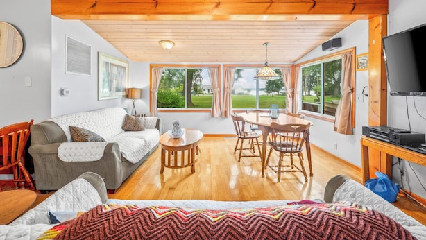 A room with a wooden ceiling, a sofa, a dining table with chairs, a TV, and large windows with curtains.
