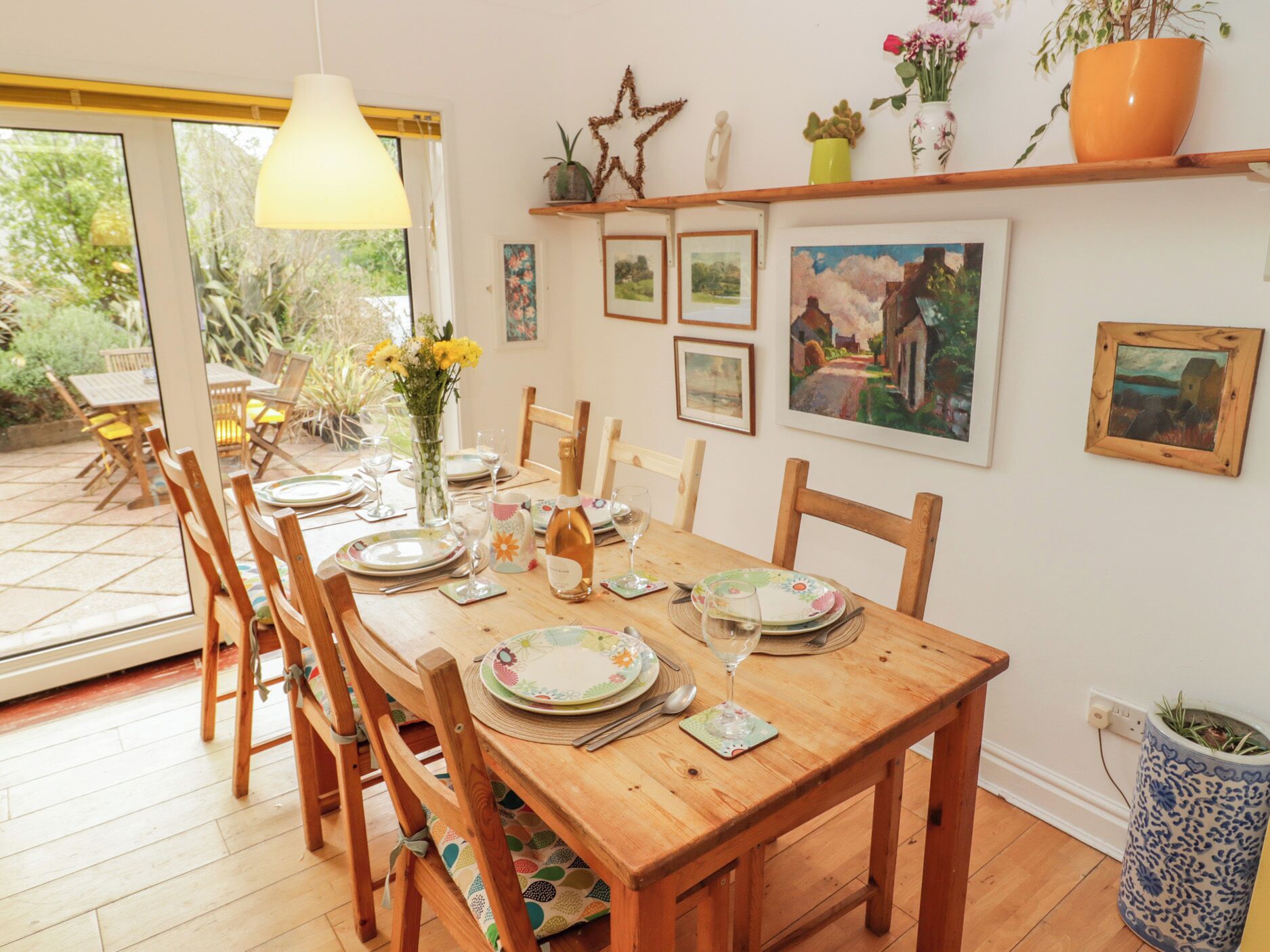 Un comedor con una mesa de madera dispuesta para seis comensales, sillas, un florero con flores y cuadros en la pared.