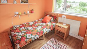Een slaapkamer met een bed, nachtkastje, lamp, raam, radiator, plank met foto's en een haardroger, plus een klein plantje.