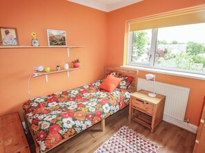 A bedroom with a bed, bedside table, lamp, window, radiator, shelf with pictures and a hairdryer, and a small plant. - 1 Rosemount Garden Villas (Tenby)
