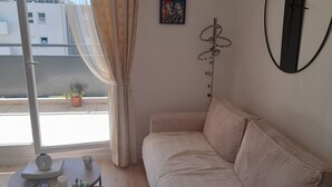 Une pièce comprenant un canapé, une petite table avec un vase et des télécommandes, une plante en pot et un appareil de climatisation.