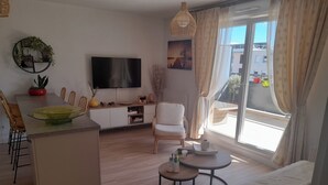 Un salon moderne doté d’un téléviseur à écran plat, d’un coin repas avec des chaises, d’un canapé et d’une petite table sur laquelle se trouvent un vase et des télécommandes.