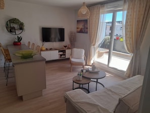 Un salon moderne doté d’un téléviseur à écran plat, d’un coin repas avec des chaises, d’un canapé et d’une petite table sur laquelle se trouvent un vase et des télécommandes.