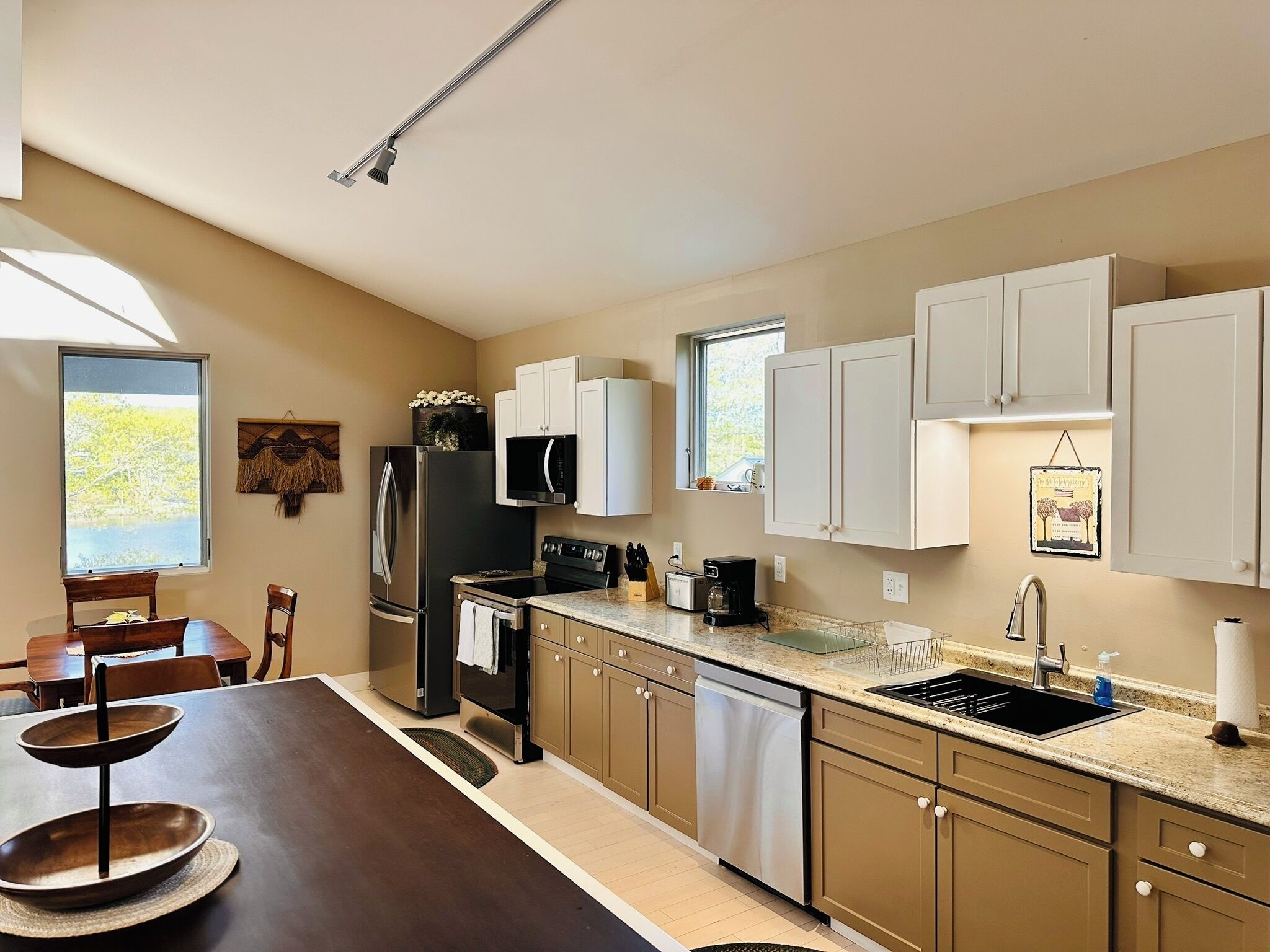 A modern kitchen with white cabinets, stainless steel appliances, a dining area with a wooden table, and a view of the outdoors.