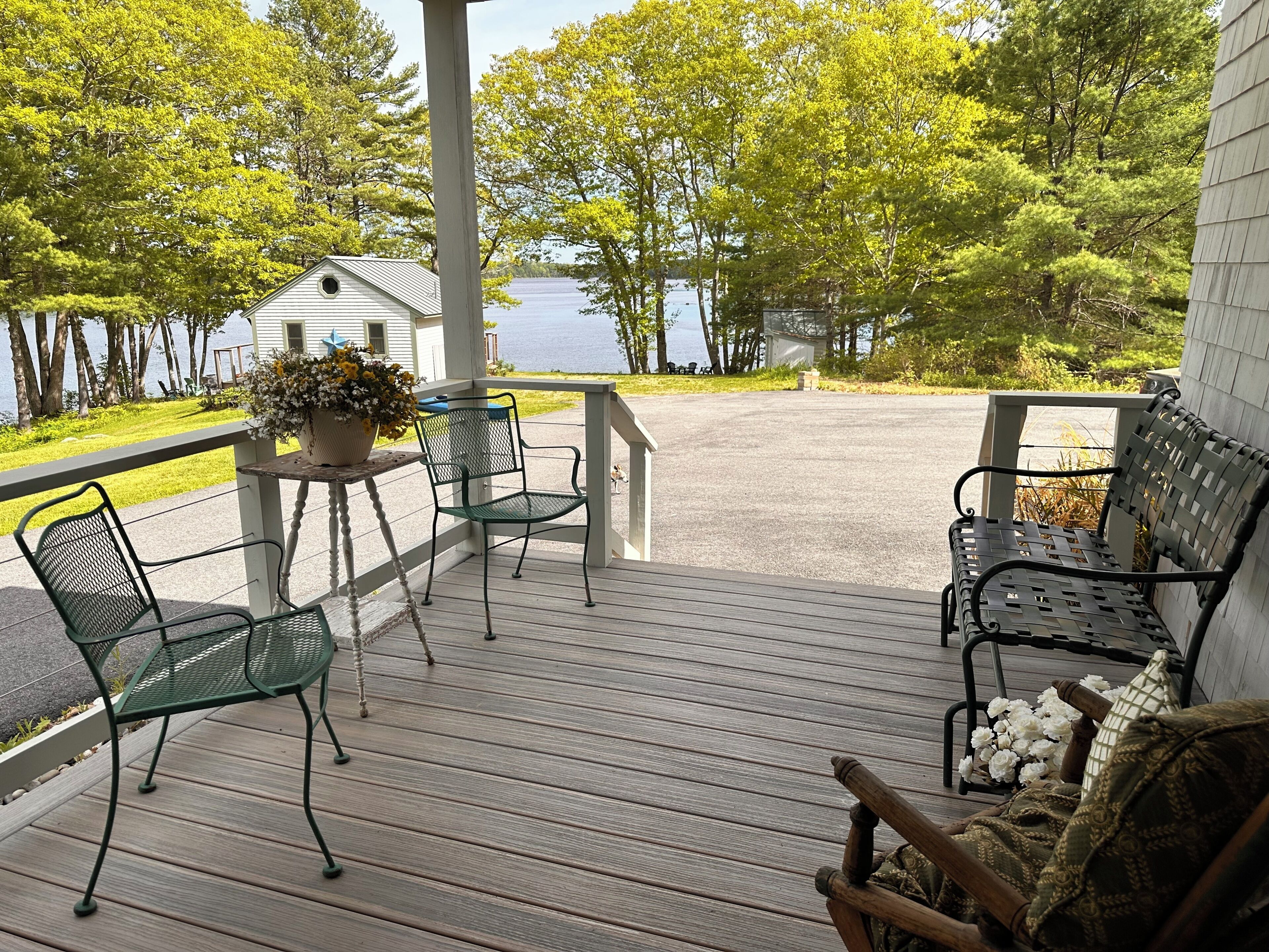 Une terrasse en bois avec des chaises en métal et un banc, donnant sur un lac bordé d’arbres.
