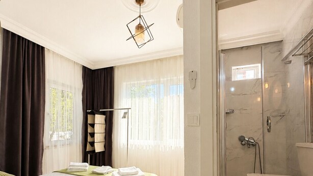 Habitación de hotel con cama, mesita de noche, zona de ducha y ventana con cortinas.