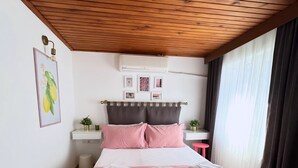 Un dormitorio ordenado con cama, mesitas de noche, un taburete pequeño y un aire acondicionado de pared.