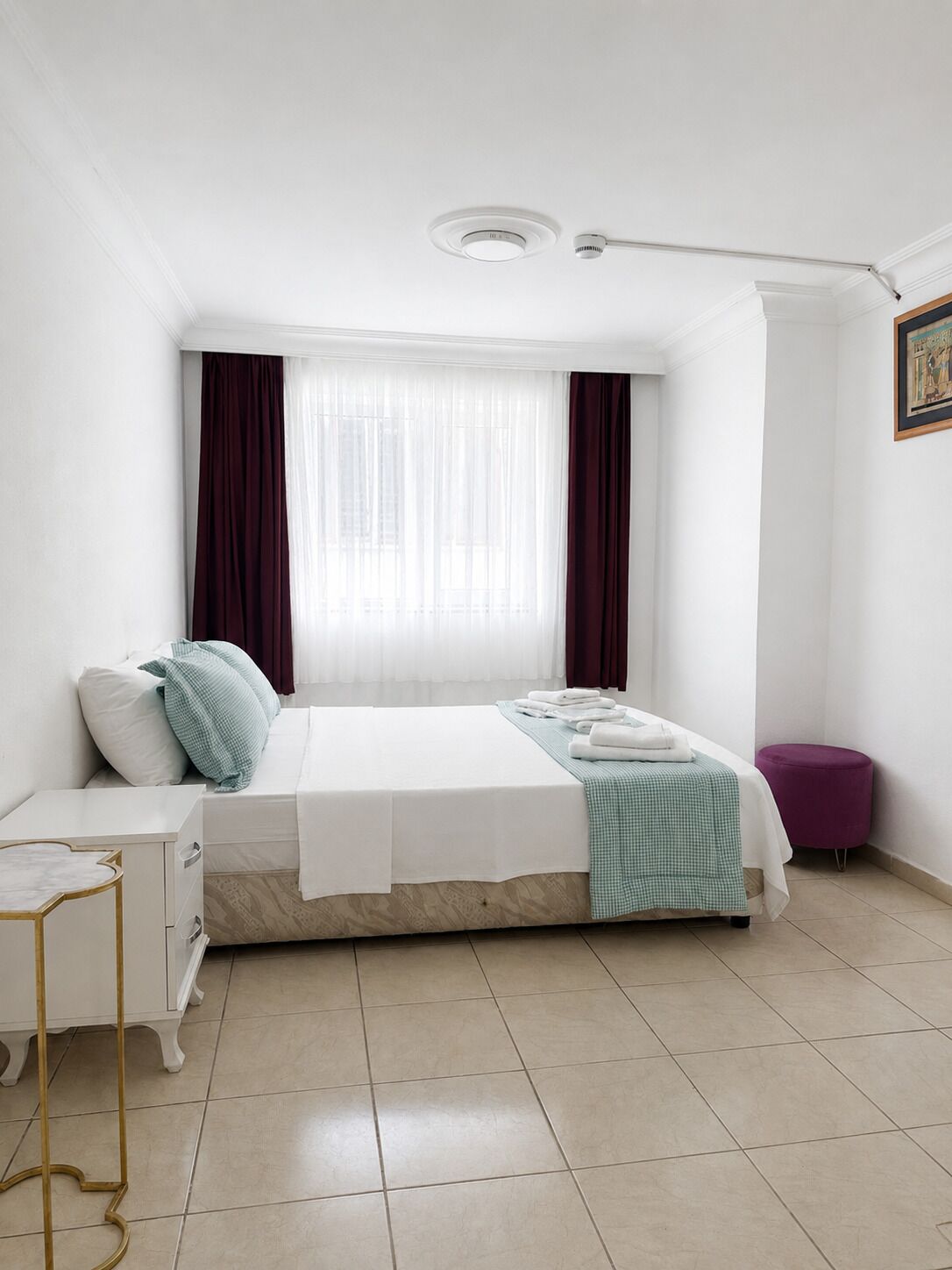 Un dormitorio con una cama, una mesita de noche, una ventana con cortinas y un taburete pequeño.