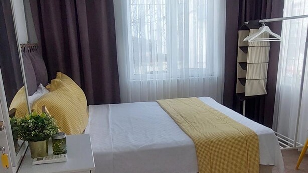 Un dormitorio con cama, mesita de noche, lámpara colgante, ventana con cortinas y armario.