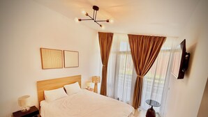 Ein Hotelzimmer mit Bett, Nachttisch, Lampe, Vorhängen, einem an der Wand montierten Fernseher und einer Deckenleuchte.