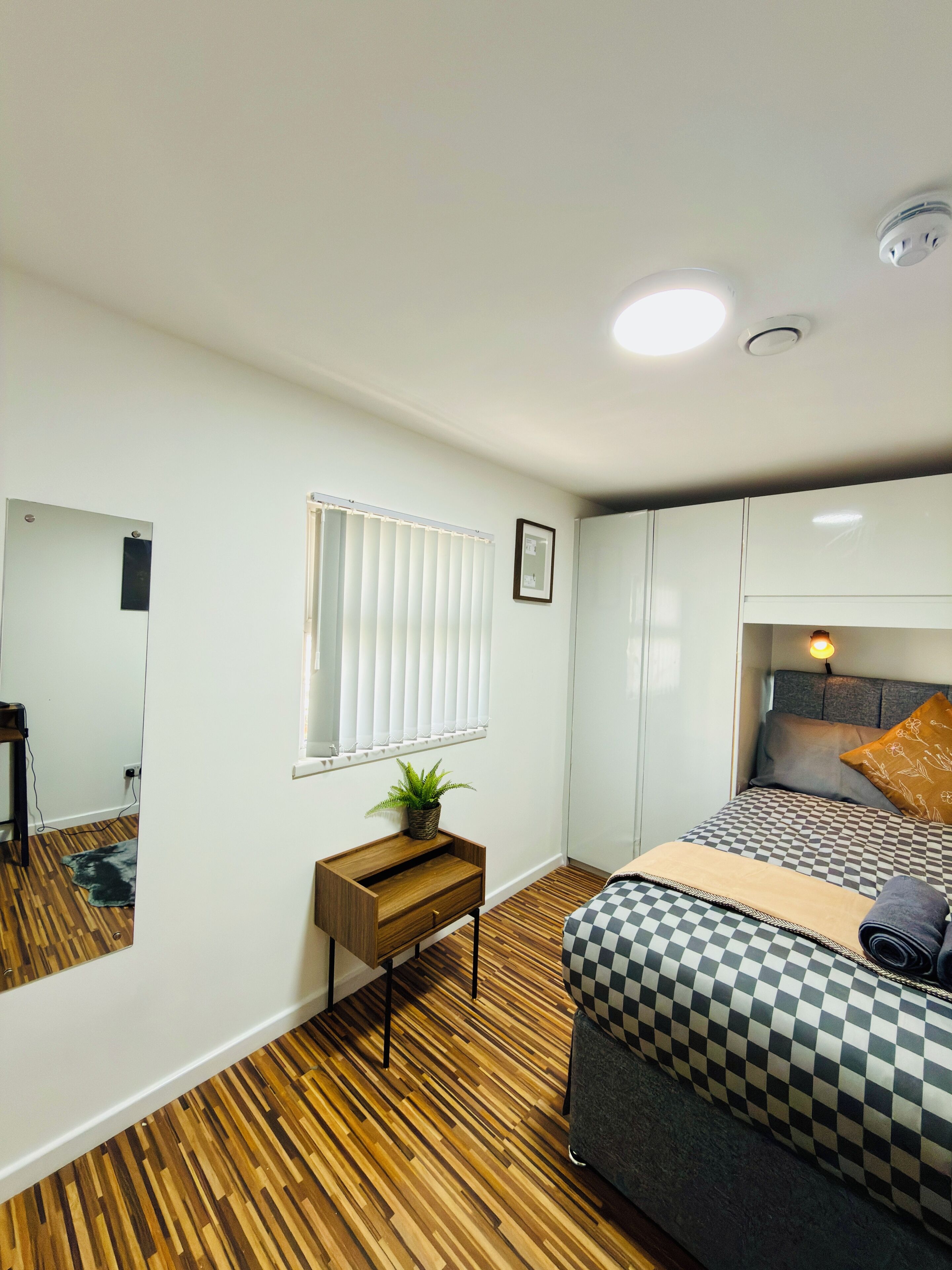 A bedroom with a bed, a bedside table with a plant, a mirror, and a window with blinds.
