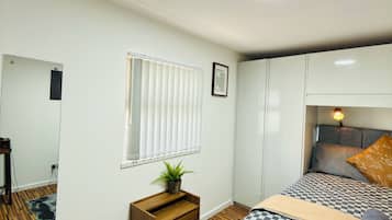 A bedroom with a bed, a bedside table with a plant, a mirror, and a window with blinds.
