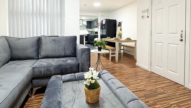 Un salon moderne avec un canapé d’angle gris, une table basse sur laquelle est posé un panier de fleurs, et une cuisine en arrière-plan.