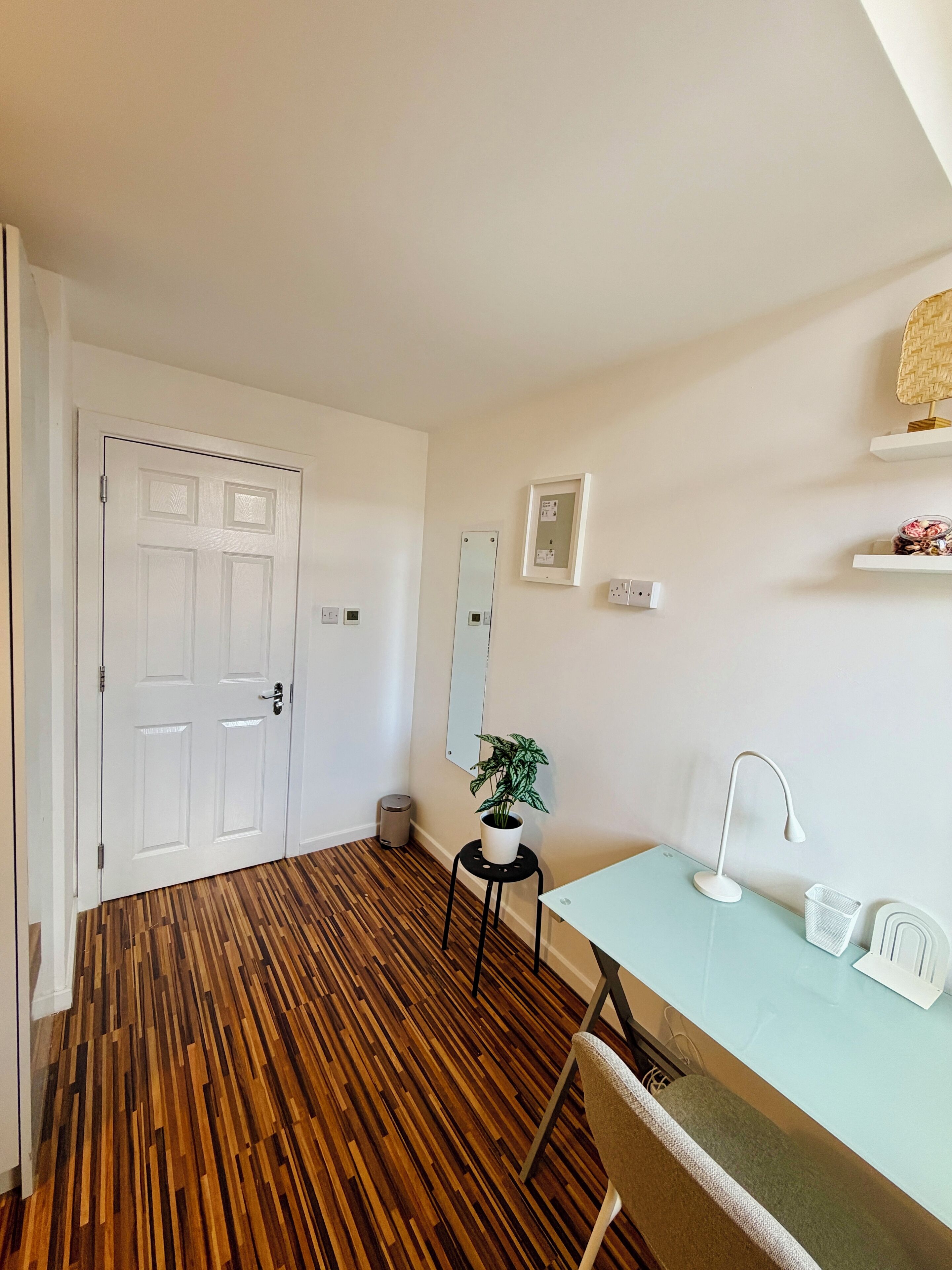 A room with a wooden floor, a white door, a small table with a chair, a desk with a lamp, a potted plant, and a mirror on the wall.