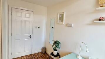A room with a wooden floor, a white door, a small table with a chair, a desk with a lamp, a potted plant, and a mirror on the wall.