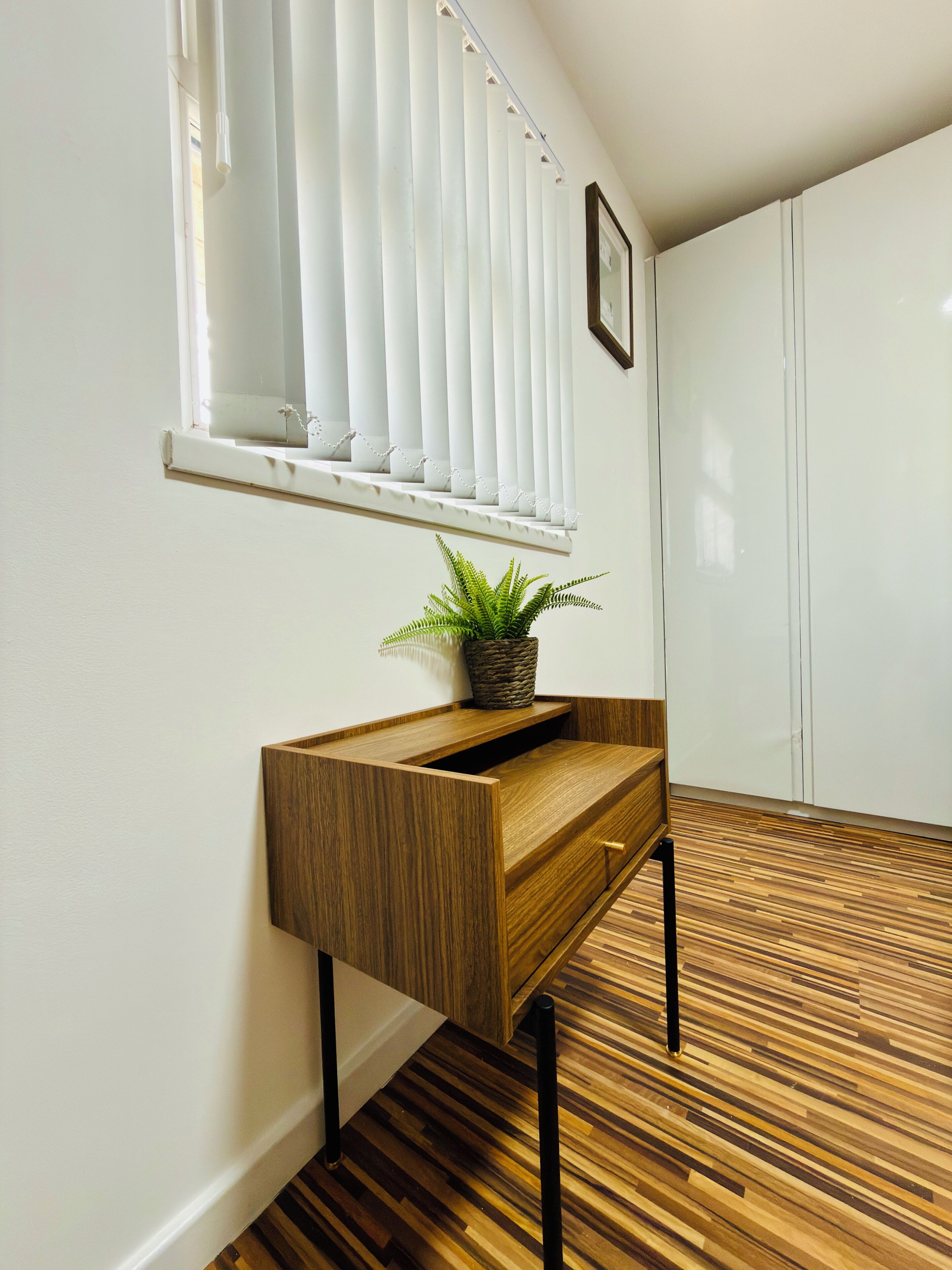 A hallway with a wooden bench, a potted plant, a window with blinds, and a wardrobe.