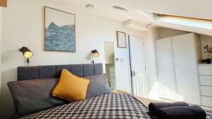 Une chambre à coucher avec une couvre-lit à carreaux, une tête de lit, deux lampes fixées au mur, une œuvre d’art encadrée et une armoire.