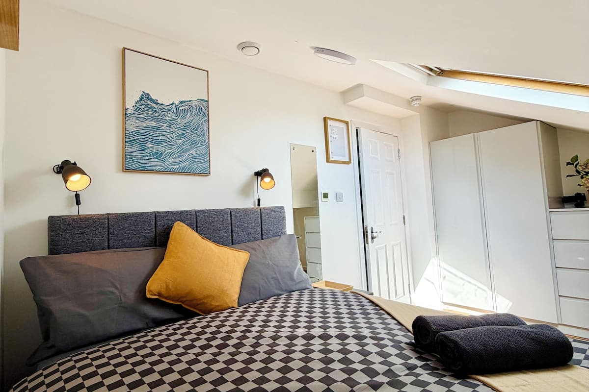 Une chambre à coucher avec une couvre-lit à carreaux, une tête de lit, deux lampes fixées au mur, une œuvre d’art encadrée et une armoire.