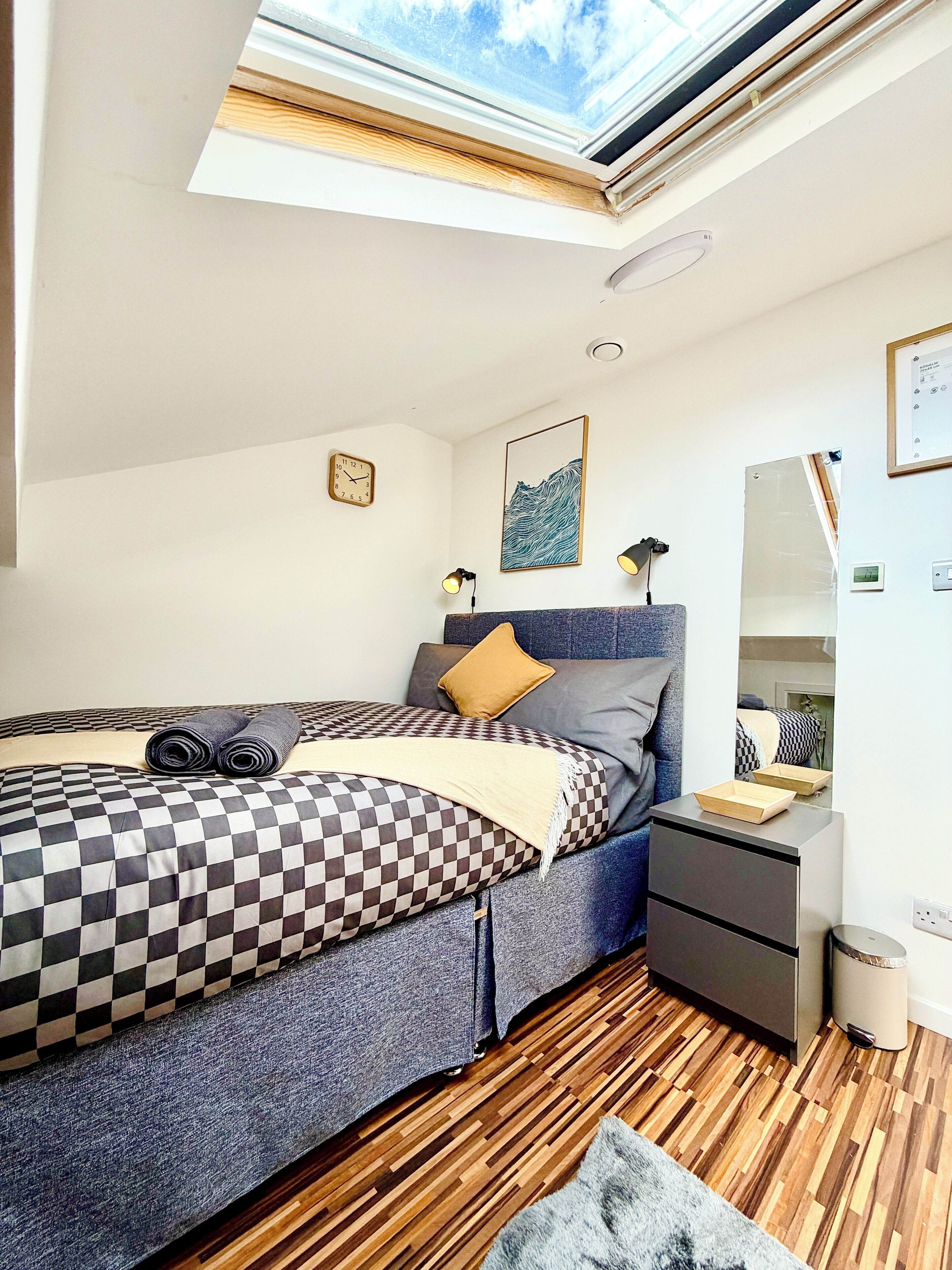 Une chambre avec une lucarne, un lit avec une couette à carreaux, une tête de lit grise, une table de chevet avec des lampes et un plancher en bois.