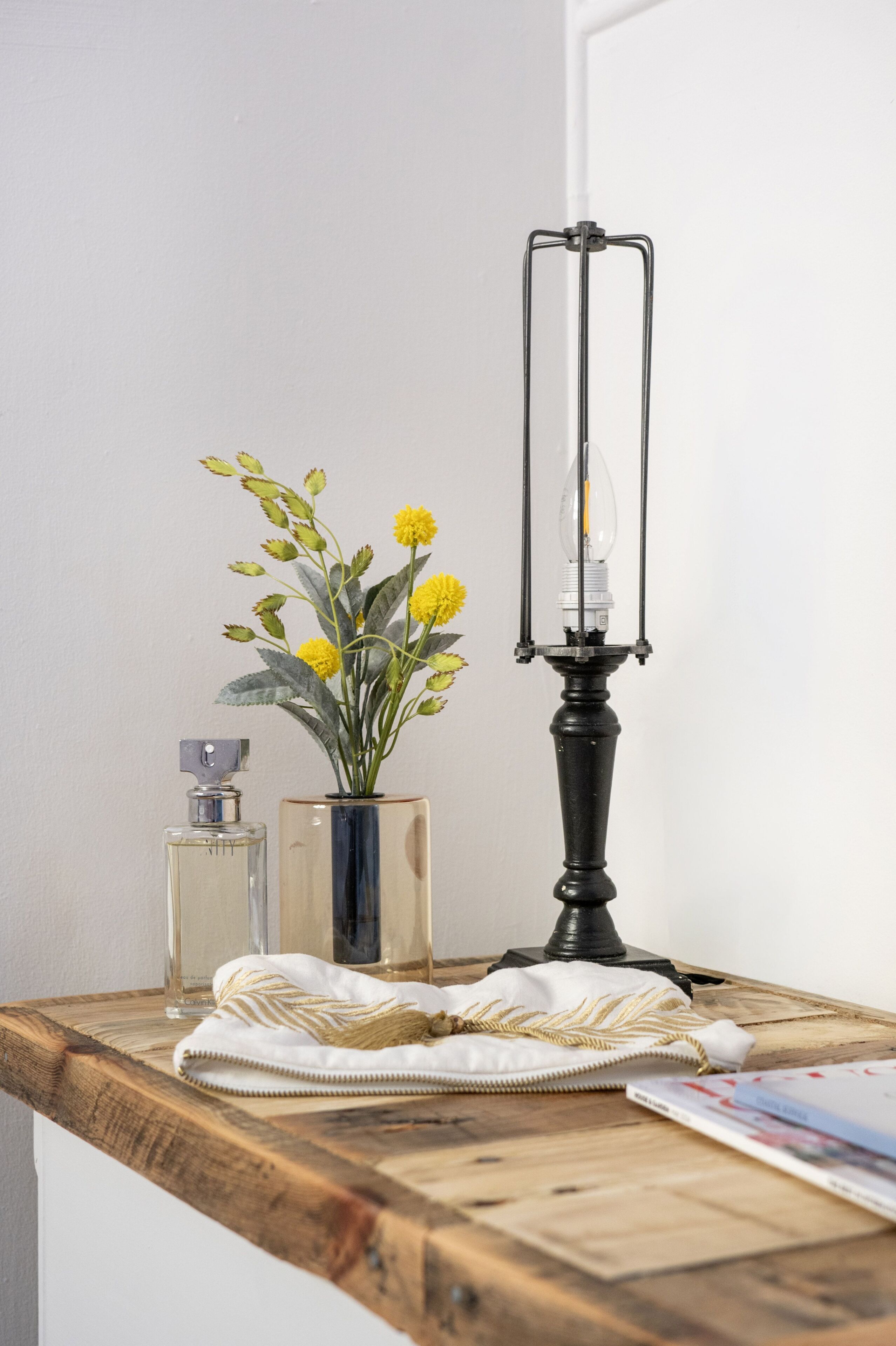 A wooden table with a lamp, a vase with yellow flowers, a perfume bottle, and a folded white cloth.