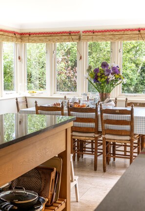 A dining area with a table set for four, surrounded by wooden chairs with cushioned seats, large windows with a view of greenery, and a wicker basket on the floor.
