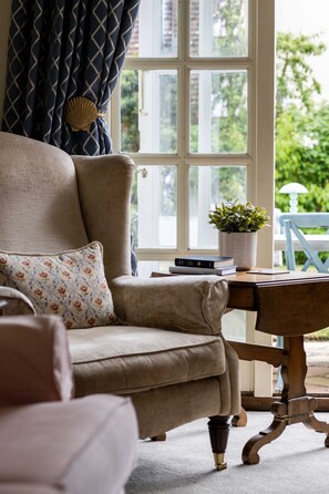 A room with a beige armchair, a wooden side table with a potted plant, and a window with a view of greenery.