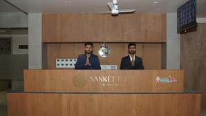A reception desk with two individuals standing behind it, a clock, and a logo for 'SANKET ELITE.' - HOTEL SANKET ELITE (Malegaon)