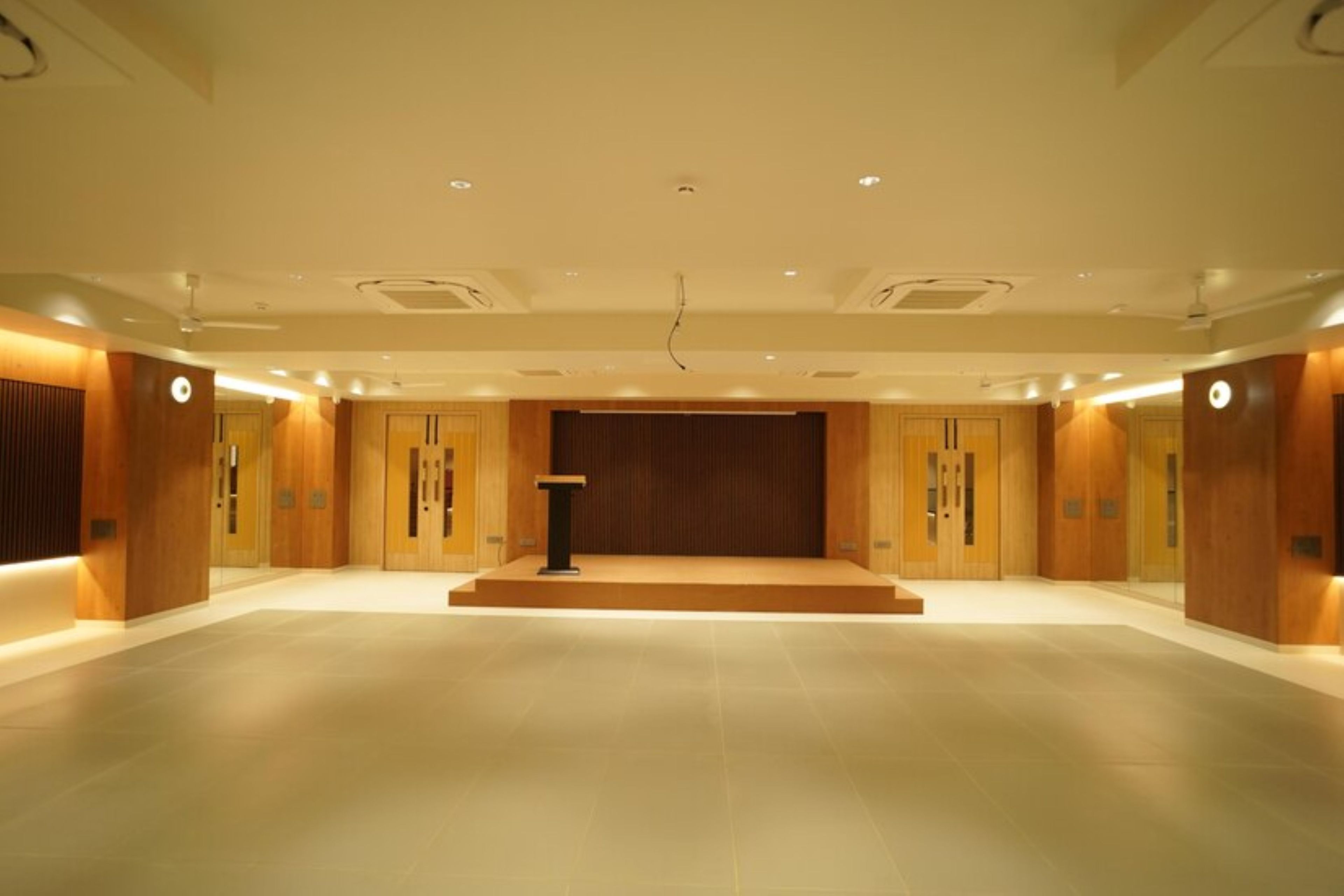 A spacious hall with a podium at the front, wooden paneling on the walls, and recessed lighting.