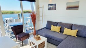 A modern living room with a grey sofa, yellow cushions, a white coffee table, and a view of a body of water through large windows. - Floating Lodge With Terrace in Lelystad (Lelystad)