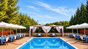 Una piscina rettangolare con bordo in mattoni, tavoli sotto ombrelloni e un pergolato in legno con tende bianche.