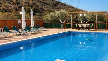 A swimming pool with lounge chairs and umbrellas, surrounded by a wooden fence and a hill in the background.
