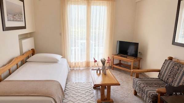 Habitación de hotel con dos camas, un televisor sobre un soporte de madera, un sofá estampado y una ventana con cortinas translúcidas.
