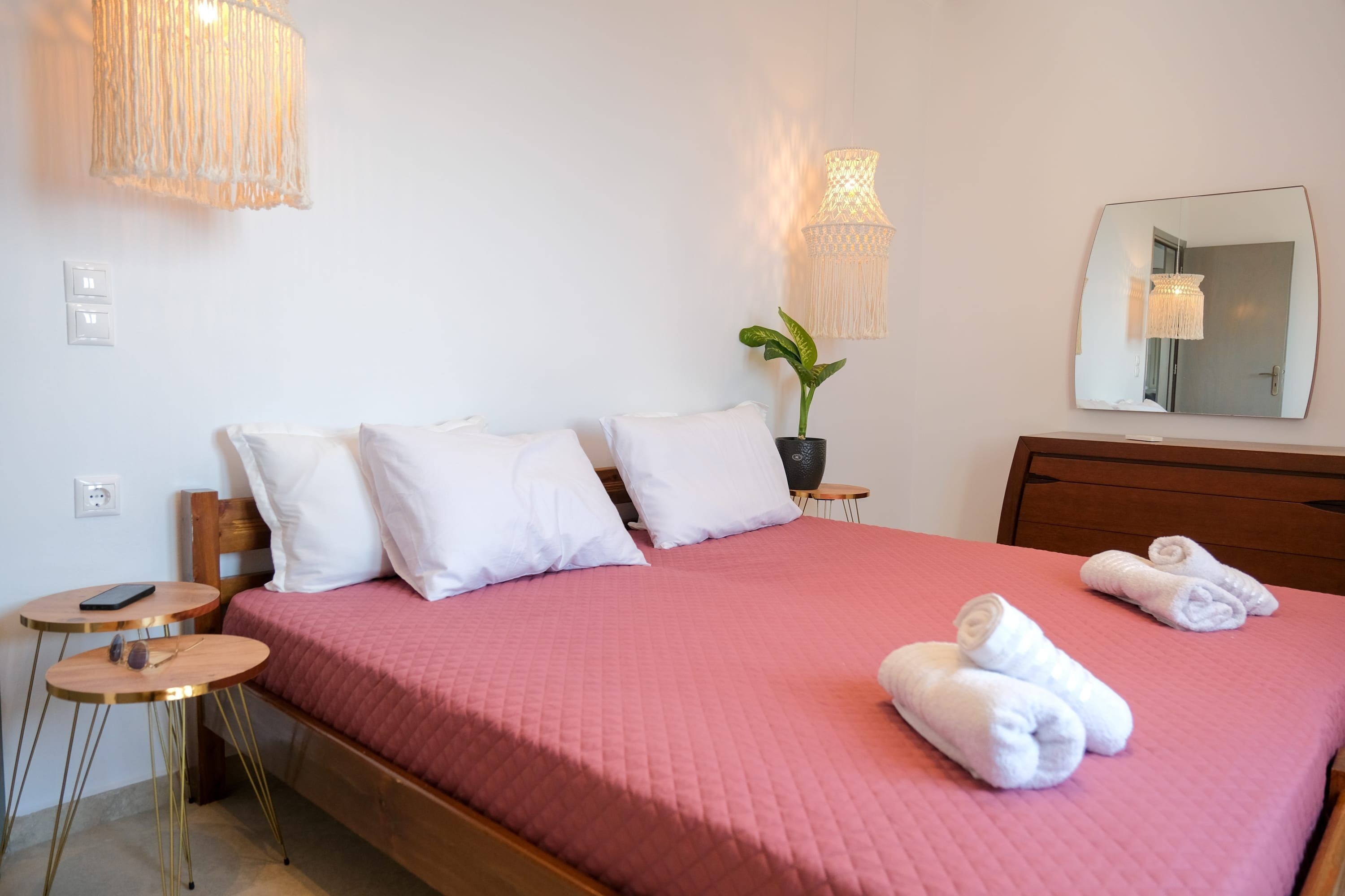 A neatly made bed with white pillows and a pink bedspread, a bedside table with a lamp, a small round table with a phone, and a potted plant.