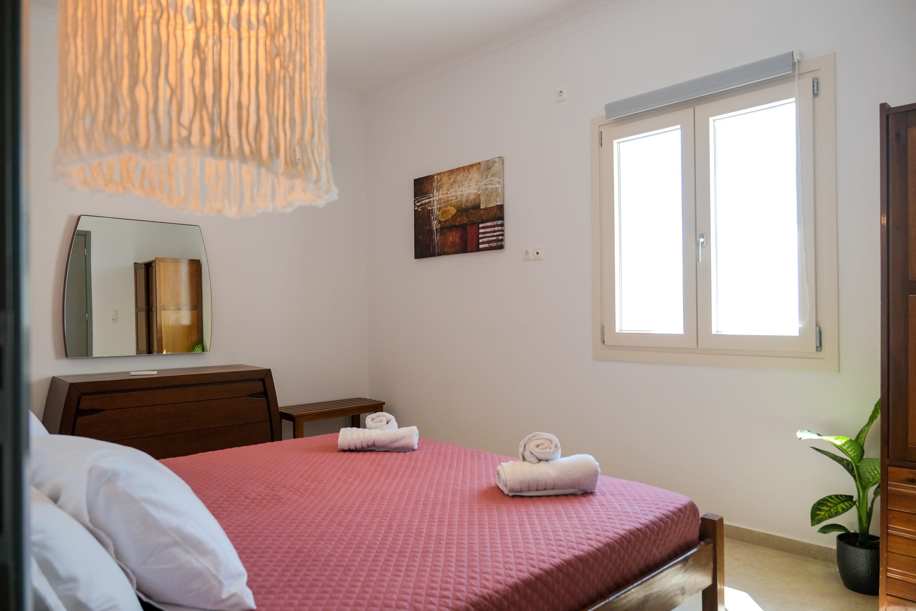 A bedroom with a bed, a mirror, a window, a painting, and a potted plant.