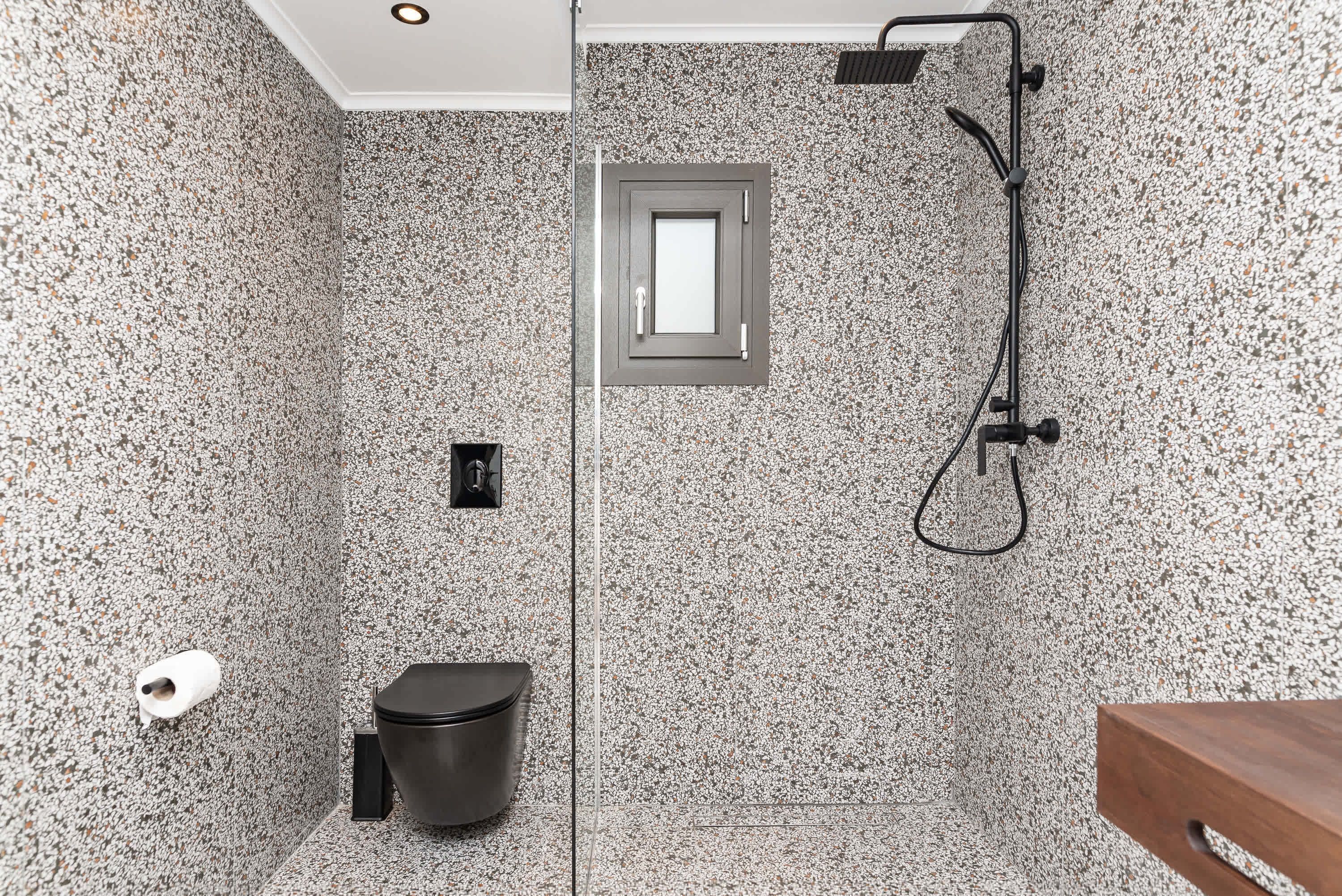 A modern bathroom with a shower, toilet, and sink, featuring speckled stone walls and a small window.