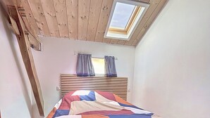 Une chambre avec un plafond en pente, un puits de lumière, un lit recouvert d’une courtepointe colorée, un plancher en bois et une fenêtre ornée de rideaux à carreaux.