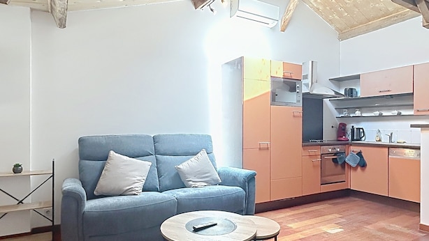 Un salon comprenant un canapé, une table basse, un plancher en bois, ainsi qu’un coin cuisine avec des placards et des appareils électroménagers.
