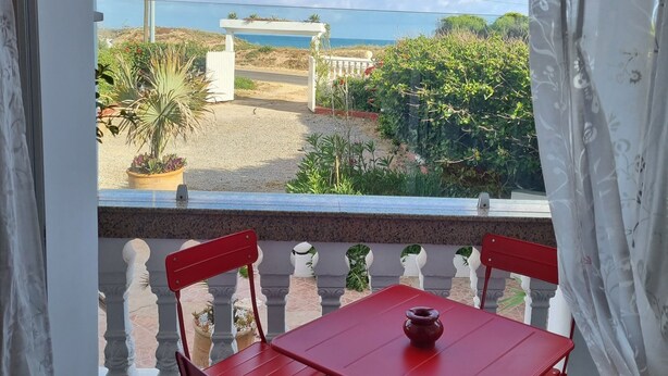 Ein Balkon mit rotem Tisch und St ühlen, Blick aufs Meer und ein Tor zu einem Garten.