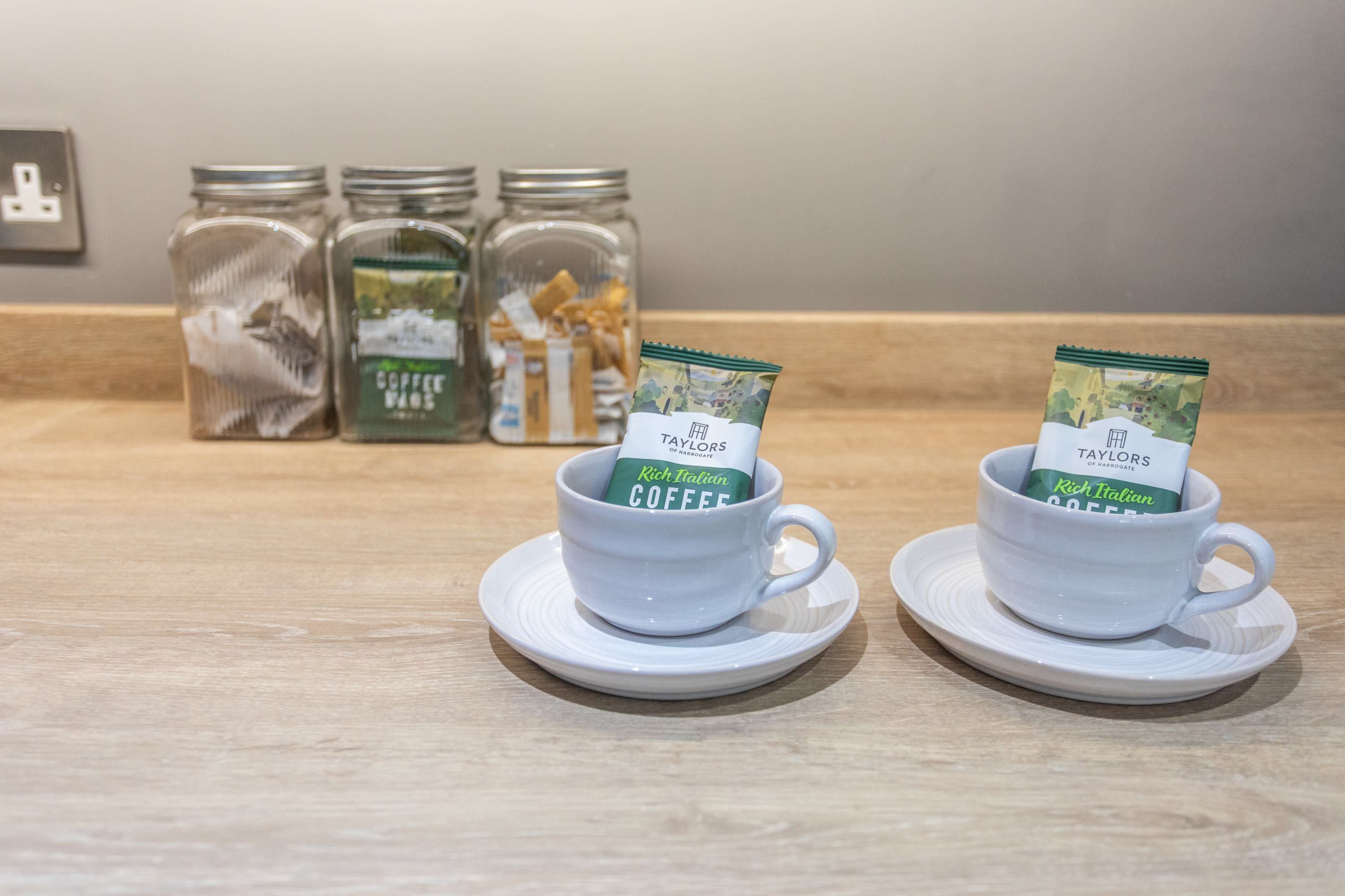 Due tazze bianche con piattini, ognuna contenente una bustina di Taylor's Rich Italian Coffee. Dietro, tre vasetti di vetro con chicchi di caffè.