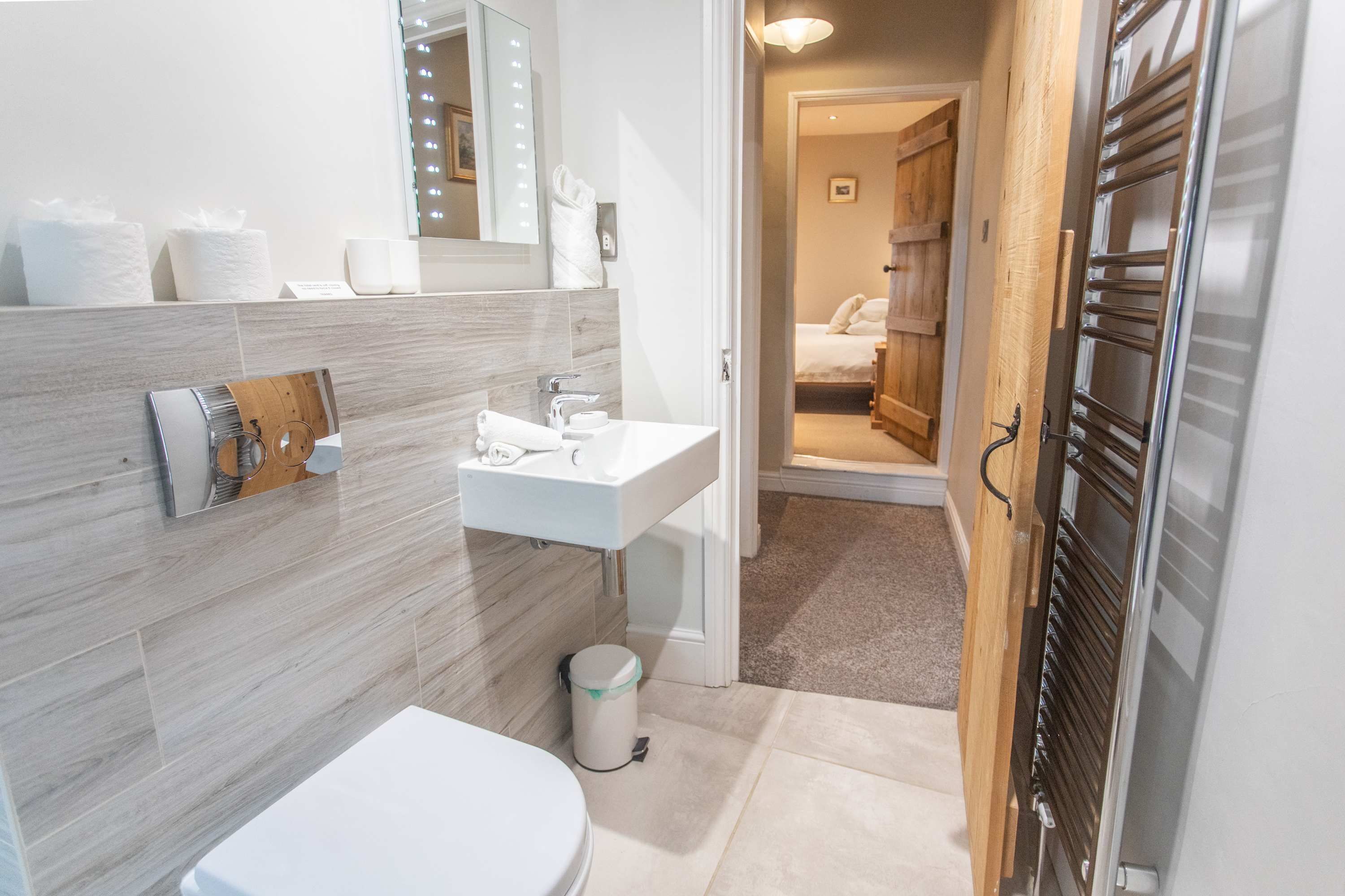 A modern bathroom with a white sink, mirror, and toilet. There is a wooden door leading to a bedroom.