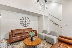 Una sala de estar con un sofá de cuero marrón, un sillón gris, una mesa de centro redonda de madera y un reloj de pared grande.