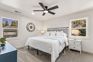 Un dormitorio con cama, mesitas de noche, ventilador de techo, reloj de pared y ventanas con vistas a árboles y edificios.